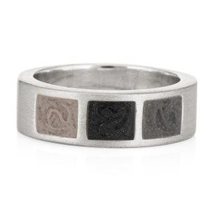 May include: A silver band ring with three square insets. The insets are made of a light brown, dark gray, and black material.