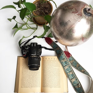Pode incluir: Uma correia de câmera vintage com um design azul-esverdeado e dourado, um globo terrestre, uma planta em vaso e um livro com as palavras "Guide to Travel" na capa.