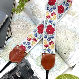 May include: A white camera strap with red, blue, and yellow floral embroidery. The strap is attached to a black camera with a large lens. The camera is lying on a map with a passport and plants in the background.