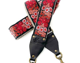 May include: A black camera strap with a red and pink floral pattern and gold hardware. The strap is made of leather and has the text "Native Stone" embossed on it.
