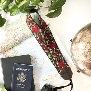 May include: A black DSLR camera with a floral patterned camera strap featuring red roses. A U.S. passport, a black notebook, and a globe are also visible, suggesting a travel theme. Green foliage surrounds the items.