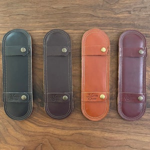 以下が含まれることがあります： 木製の表面に配置された、さまざまな茶色の革製ペンケース4つ。各ケースには真鍮製のボタンが付いたストラップ留め具が付いています。「Native Sons」の文字がケースの下部に印刷されています。