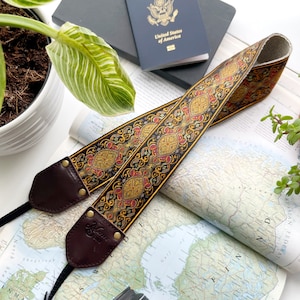 May include: An intricately patterned camera strap in black, gold, and red, lying on a map. A passport and a camera lens are also visible. The strap features brown leather ends and the brand name "Laziz Tone" is printed on the leather.