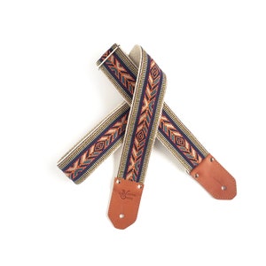 Puede incluir: Una correa de guitarra con un patr&oacute;n tejido en tonos naranja, azul, verde y blanco. La correa tiene extremos de cuero marr&oacute;n con el texto "Native 0392" grabado en un extremo.