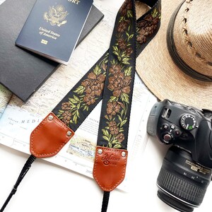 Può includere: Tracolla per fotocamera nera con ricami floreali marroni e estremità in pelle. La tracolla presenta il testo "NATIVE SONS" alle estremità in pelle. Un passaporto, una mappa, un cappello e una fotocamera sono presenti nell'immagine.