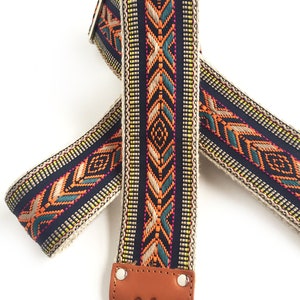 Puede incluir: Una correa de guitarra con un dise&ntilde;o tejido en tonos de azul, naranja, verde y amarillo. La correa tiene un extremo de cuero marr&oacute;n con el texto "Native Sons" grabado.