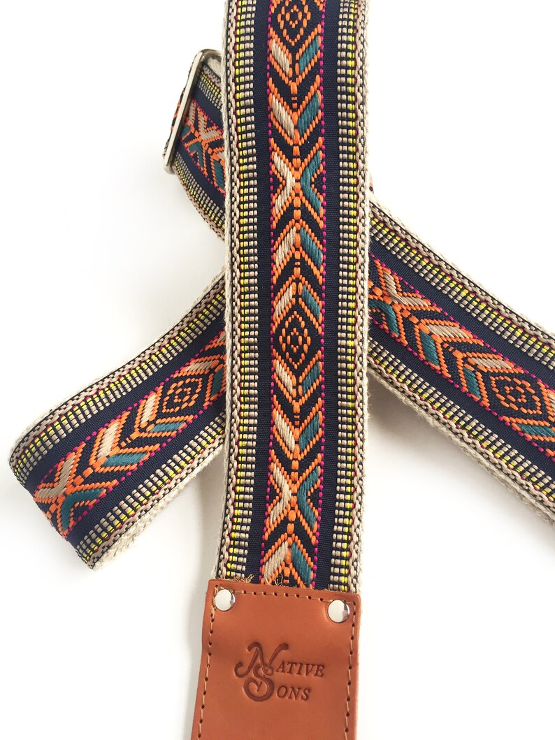 Puede incluir: Una correa de guitarra con un dise&ntilde;o tejido en tonos de azul, naranja, verde y amarillo. La correa tiene un extremo de cuero marr&oacute;n con el texto "Native Sons" grabado.