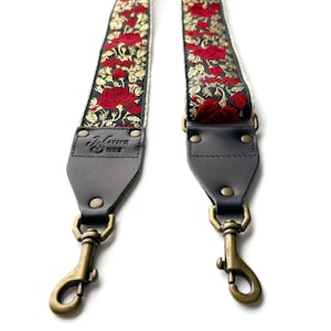 May include: A black and white guitar strap with red embroidered roses and gold accents. The strap has leather ends with brass clasps and a black leather patch with the text "Native One" in white.