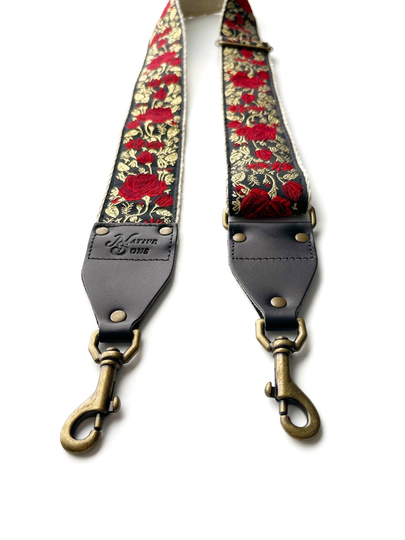 May include: A black and white guitar strap with red embroidered roses and gold accents. The strap has leather ends with brass clasps and a black leather patch with the text "Native One" in white.