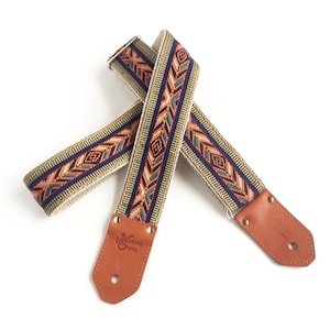 Puede incluir: Una correa de guitarra con un patr&oacute;n geom&eacute;trico colorido y extremos de cuero marr&oacute;n. La correa est&aacute; hecha por Native Sons.