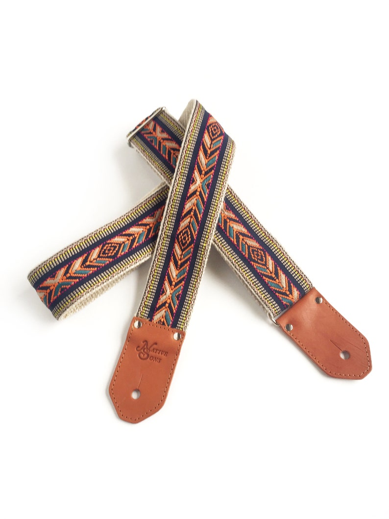 Puede incluir: Una correa de guitarra con un patr&oacute;n geom&eacute;trico colorido y extremos de cuero marr&oacute;n. La correa est&aacute; hecha por Native Sons.
