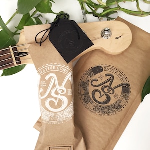 Puede incluir: Una bolsa de papel marr&oacute;n con un logotipo blanco que dice "Native Sons" y "Play in Style" con un dise&ntilde;o floral. La bolsa est&aacute; etiquetada como "Guitar Strap" y tiene una etiqueta negra con el mismo logotipo en negro.