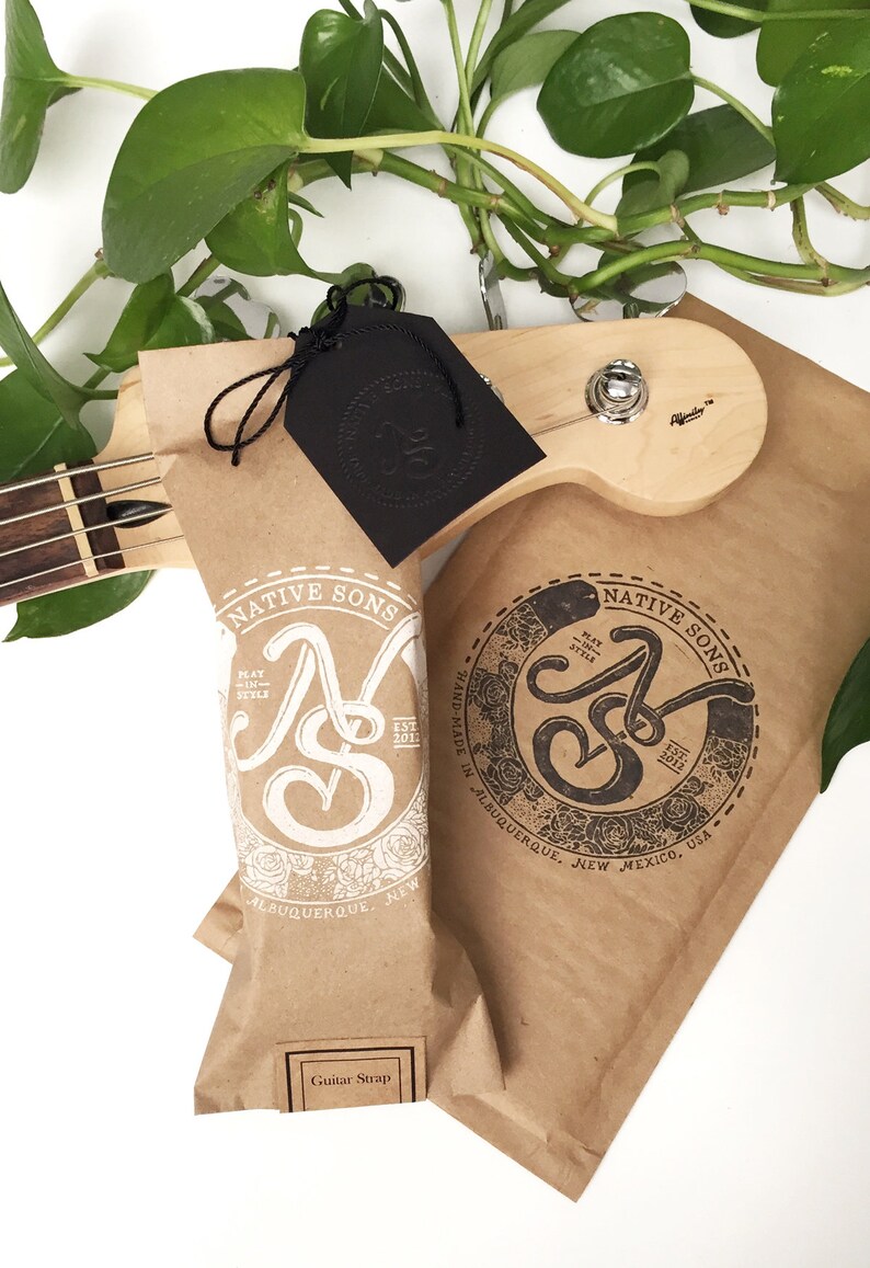 Puede incluir: Una bolsa de papel marr&oacute;n con un logotipo blanco que dice "Native Sons" y "Play in Style" con un dise&ntilde;o floral. La bolsa est&aacute; etiquetada como "Guitar Strap" y tiene una etiqueta negra con el mismo logotipo en negro.