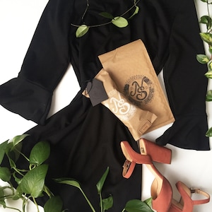 May include: A black dress with long sleeves and a bell bottom hemline. The dress is laid out on a white surface with a brown paper bag and a pair of coral colored heeled sandals. Green leafy vines are draped over the dress and sandals.