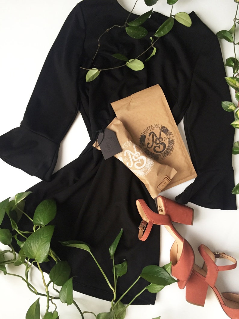 May include: A black dress with long sleeves and a bell bottom hemline. The dress is laid out on a white surface with a brown paper bag and a pair of coral colored heeled sandals. Green leafy vines are draped over the dress and sandals.