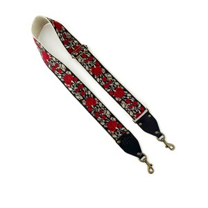 May include: A black camera strap with red embroidered roses and gold accents. The strap has two adjustable metal clasps.