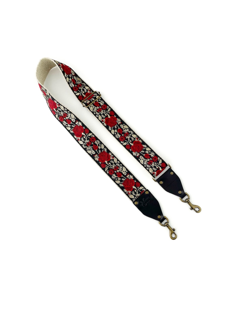May include: A black camera strap with red embroidered roses and gold accents. The strap has two adjustable metal clasps.
