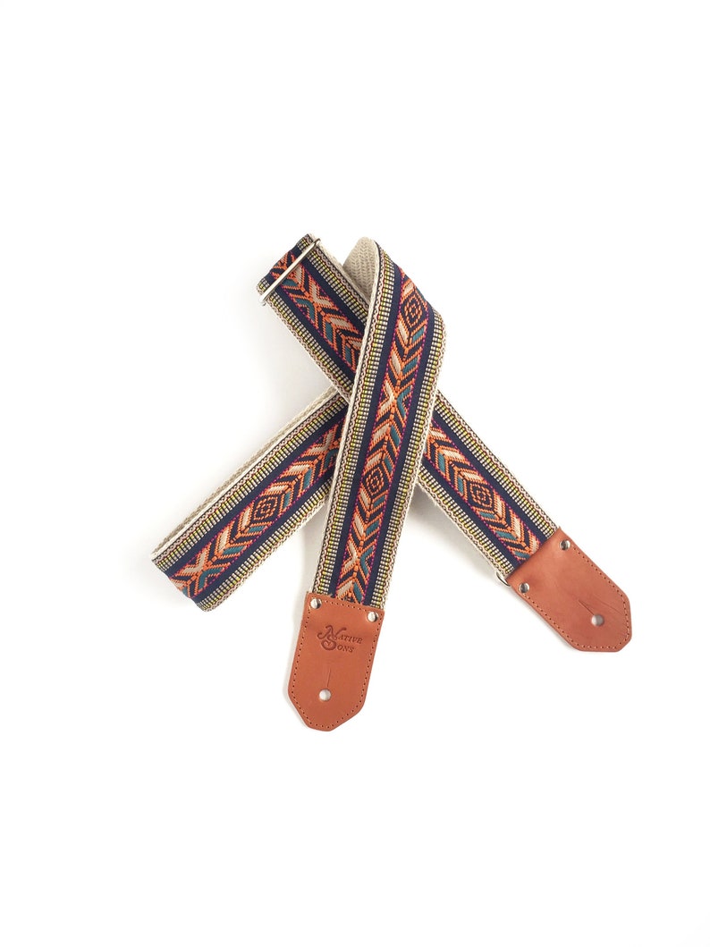 Puede incluir: Una correa de guitarra con un patr&oacute;n tejido en tonos naranja, azul, verde y blanco. La correa tiene extremos de cuero marr&oacute;n con el texto "Native 0392" grabado en un extremo.
