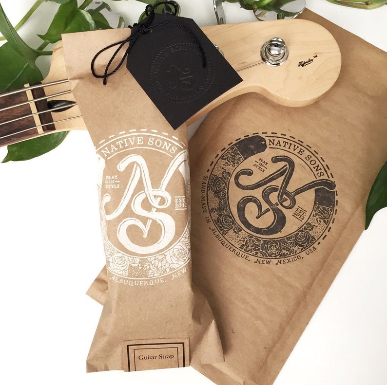 May include: A brown paper bag with a white logo that says "Native Sons" and "Play in Style" with a white floral design. The bag is labeled "Guitar Strap" and has a black tag with a logo that says "Native Sons".