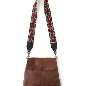 May include: A brown leather crossbody bag with a black and red floral patterned strap. The strap has gold hardware.