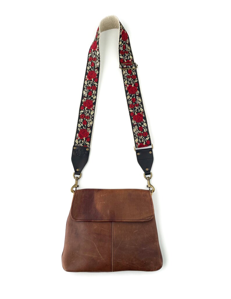 May include: A brown leather crossbody bag with a black and red floral patterned strap. The strap has gold hardware.