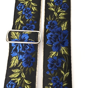 May include: A black guitar strap with a floral pattern featuring blue and green flowers. The strap has a silver buckle.