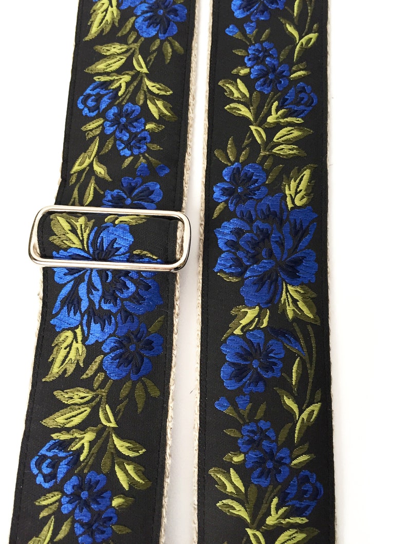 May include: A black guitar strap with a floral pattern featuring blue and green flowers. The strap has a silver buckle.