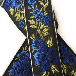 May include: A black guitar strap with a blue and green floral pattern. The strap is made of a woven fabric with a natural-colored edge.