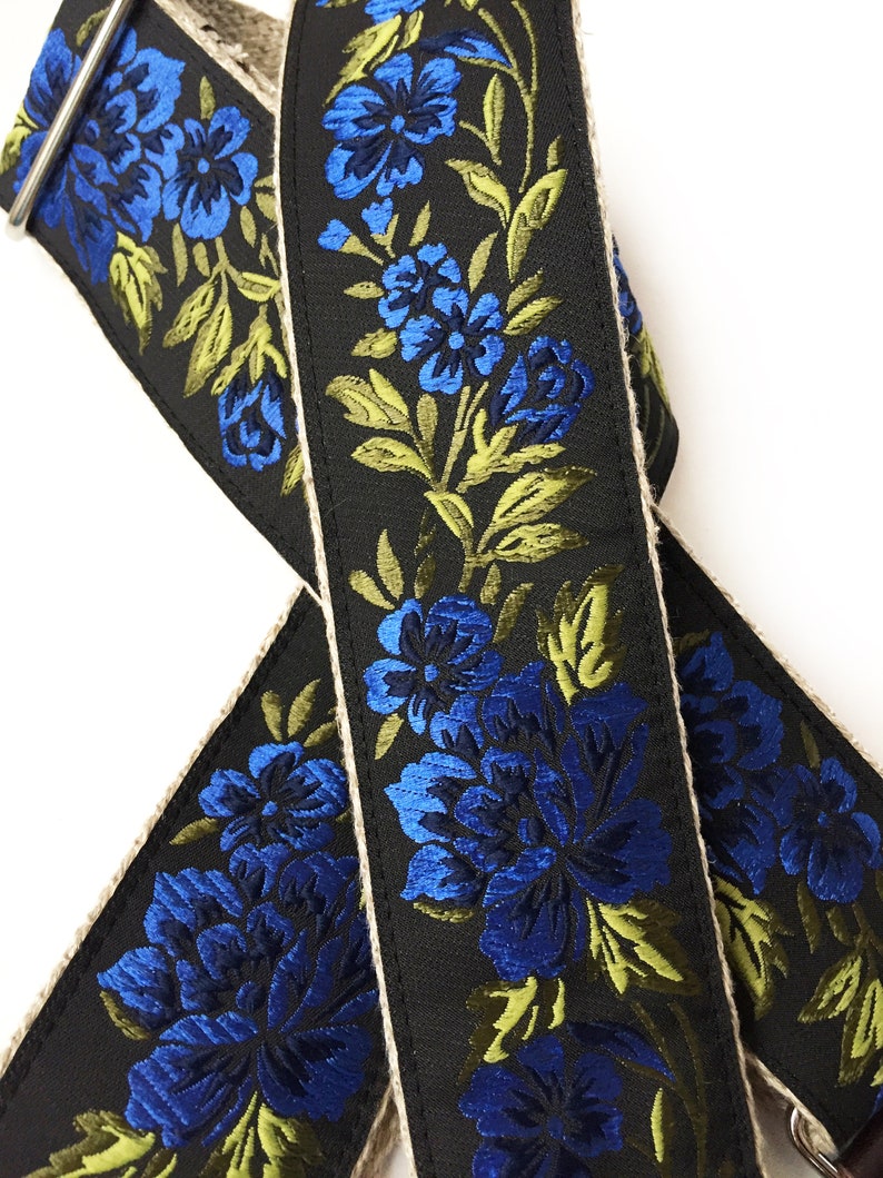May include: A black guitar strap with a blue and green floral pattern. The strap is made of a woven fabric with a natural-colored edge.