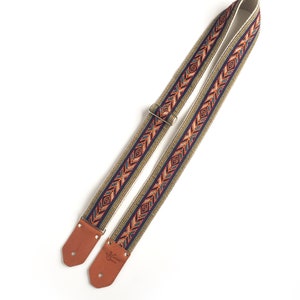 Puede incluir: Una correa de guitarra con una base beige y un patr&oacute;n geom&eacute;trico colorido en tonos naranja, azul y verde. La correa tiene extremos de cuero marr&oacute;n con el texto "Genuine Leather" grabado en un extremo.