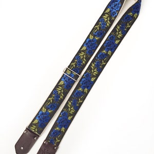 May include: A black guitar strap with blue and green embroidered flowers. The strap has brown leather ends with a silver buckle.