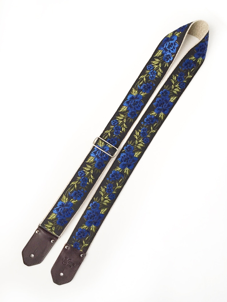 May include: A black guitar strap with blue and green embroidered flowers. The strap has brown leather ends with a silver buckle.