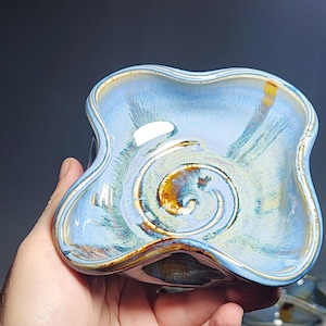 May include: A handmade ceramic bowl with a unique, wavy shape and a light blue glaze. The interior features a spiral design with brown and gold accents. The bowl is held in a hand, showcasing its size and form.