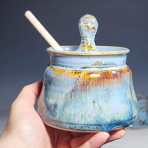 May include: A handmade ceramic jar with a lid and a wooden spoon. The jar is a light blue with brown and yellow accents. The jar is held in a hand.