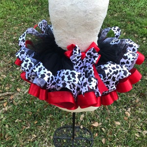 May include: A black tulle tutu with a white and black cow print ribbon and red satin ribbon. The tutu is on a white dress form.