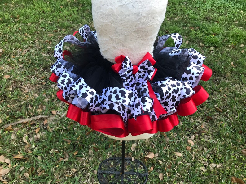 May include: A black tulle tutu with a white and black cow print ribbon and red satin ribbon. The tutu is on a white dress form.