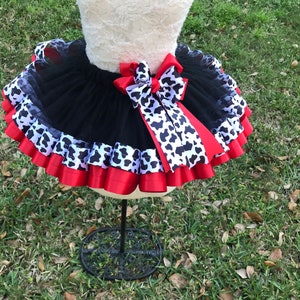 May include: Black tulle tutu skirt with red and black and white cow print ribbon. The skirt has multiple layers of tulle and is decorated with a large red bow.