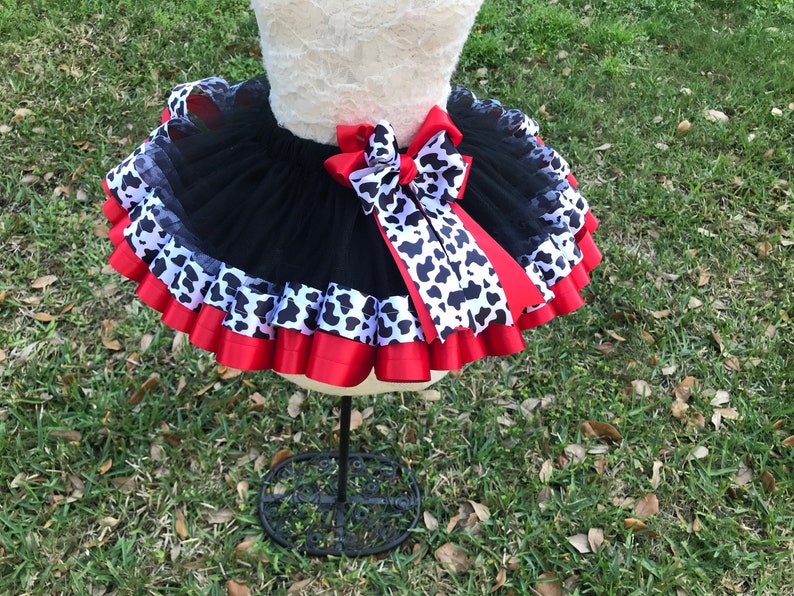 May include: Black tulle tutu skirt with red and black and white cow print ribbon. The skirt has multiple layers of tulle and is decorated with a large red bow.