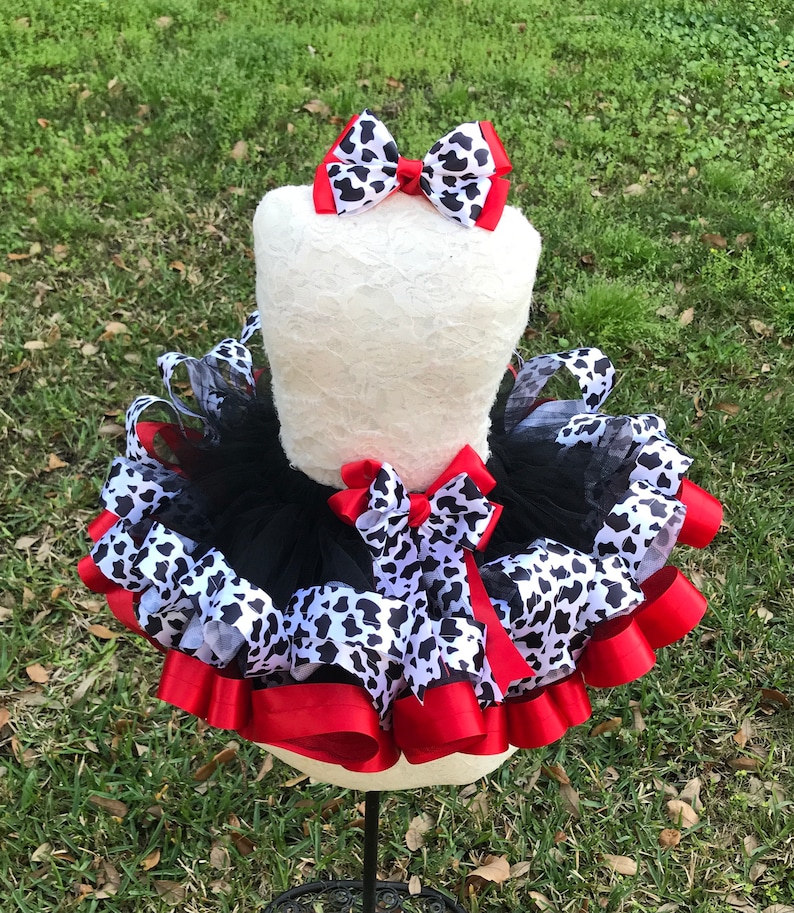 May include: A black tulle tutu with red and black and white cow print ribbon accents. A matching cow print bow is on top of the tutu.