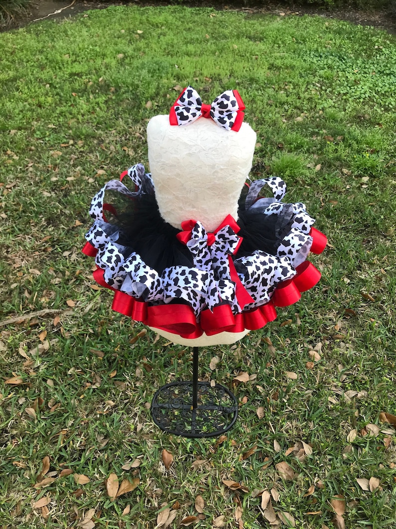 May include: A black tulle tutu skirt with red and white cow print ribbon accents. The skirt is on a white dress form and has a red bow on top.