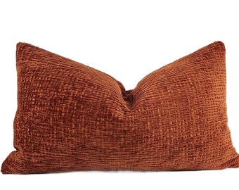 May include: A rectangular throw pillow in a deep burnt orange color. The fabric features a distinct, raised, and textured grid-like pattern across the entire surface, creating a tactile appearance with subtle variations in light and shadow.