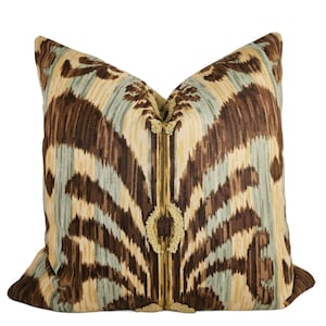 May include: Decorative throw pillow with a brown, tan, and light blue ikat pattern. The pillow has a vertical gold trim with a decorative circular element. The pillow is square and has a soft, textured appearance.
