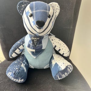 K&ouml;nnte beinhalten: Ein handgefertigter Teddyb&auml;r mit einem Patchwork-Design in Blau- und Wei&szlig;t&ouml;nen. Der B&auml;r hat Sternmuster, Karomuster und Jeansstoffe. Der B&auml;r hat den Namen "Grayson" auf einem Fu&szlig; und die Zahlen "6-22-23" auf dem anderen gestickt. Der B&auml;r hat auch ein kleines Oshkosh B'gosh-Etikett.