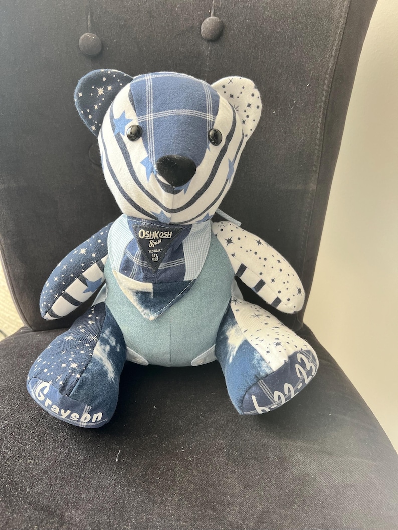 K&ouml;nnte beinhalten: Ein handgefertigter Teddyb&auml;r mit einem Patchwork-Design in Blau- und Wei&szlig;t&ouml;nen. Der B&auml;r hat Sternmuster, Karomuster und Jeansstoffe. Der B&auml;r hat den Namen "Grayson" auf einem Fu&szlig; und die Zahlen "6-22-23" auf dem anderen gestickt. Der B&auml;r hat auch ein kleines Oshkosh B'gosh-Etikett.