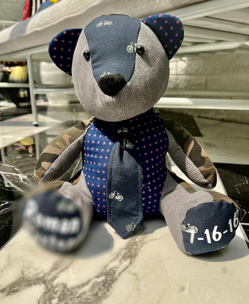 K&ouml;nnte beinhalten: Ein handgefertigter Teddyb&auml;r im Patchwork-Design. Der B&auml;r hat einen grauen K&ouml;rper, marineblaue Akzente mit Fahrradmustern und Tarnfarbenarme. Der B&auml;r hat eine marineblaue Krawatte und das Datum "7-16-16" auf einem Fu&szlig;.