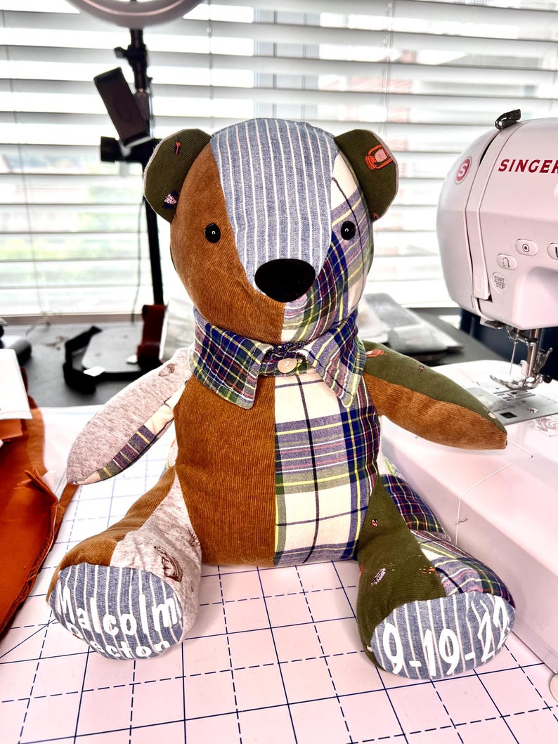 K&ouml;nnte beinhalten: Ein Patchwork-Teddyb&auml;r mit einem braunen Cord-K&ouml;rper, karierten und gestreiften Stoffakzenten und einer schwarzen Nase. Der B&auml;r hat einen karierten Kragen und der Name "Malcolm" und das Datum "9-19-22" sind auf seine F&uuml;&szlig;e gedruckt. Im Hintergrund ist eine Singer-N&auml;hmaschine zu sehen.