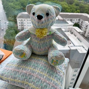K&ouml;nnte beinhalten: Ein handgefertigter Teddyb&auml;r mit Patchwork-Design in Pastellfarben, der auf einem passenden Kissen sitzt. Der B&auml;r hat einen gelben Blumenkragen und der Name "Paisleigh" und das Datum "4-5-25" sind auf dem B&auml;ren und Kissen gestickt.