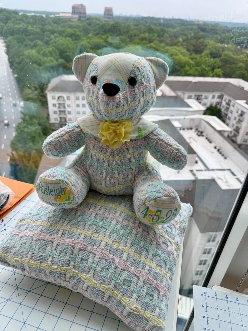 K&ouml;nnte beinhalten: Ein handgefertigter Teddyb&auml;r mit Patchwork-Design in Pastellfarben, der auf einem passenden Kissen sitzt. Der B&auml;r hat einen gelben Blumenkragen und der Name "Paisleigh" und das Datum "4-5-25" sind auf dem B&auml;ren und Kissen gestickt.