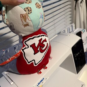 K&ouml;nnte beinhalten: Ein handgefertigter Stoffb&auml;r mit Patchwork-Design. Der B&auml;renkopf hat einen hellblauen Stoff mit Keks-Mustern, und der K&ouml;rper hat einen rot-wei&szlig;en Aufn&auml;her mit einem "KC"-Logo. Der B&auml;r liegt auf einer wei&szlig;en N&auml;hmaschine.