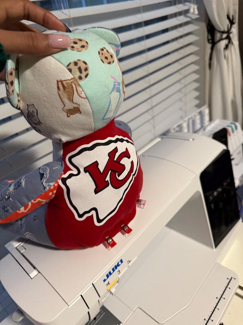 K&ouml;nnte beinhalten: Ein handgefertigter Stoffb&auml;r mit Patchwork-Design. Der B&auml;renkopf hat einen hellblauen Stoff mit Keks-Mustern, und der K&ouml;rper hat einen rot-wei&szlig;en Aufn&auml;her mit einem "KC"-Logo. Der B&auml;r liegt auf einer wei&szlig;en N&auml;hmaschine.